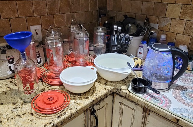 Kitchen counter set up for making hummingbird nectar, with multiple red hummingbird feeders, bowls, measuring tools, and an electric kettle used to boil water.