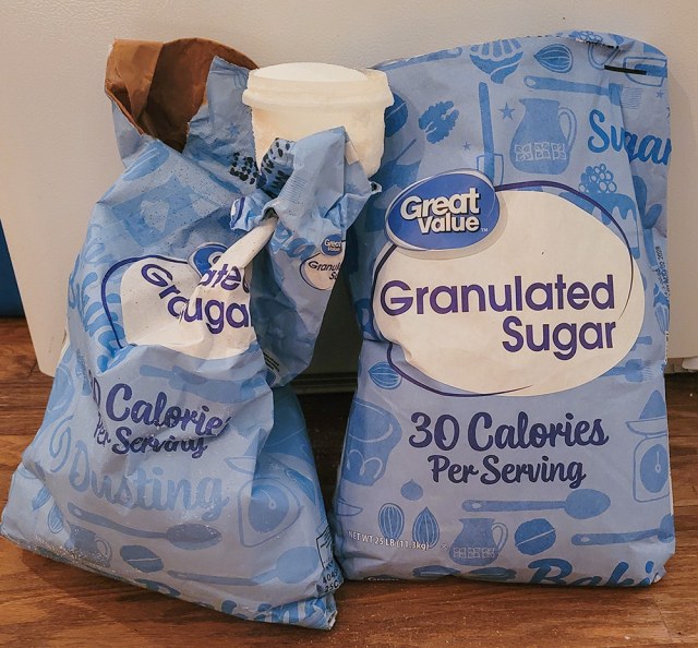 Two large 25-pound bags of granulated white sugar standing side by side, one partly used and one unopened.