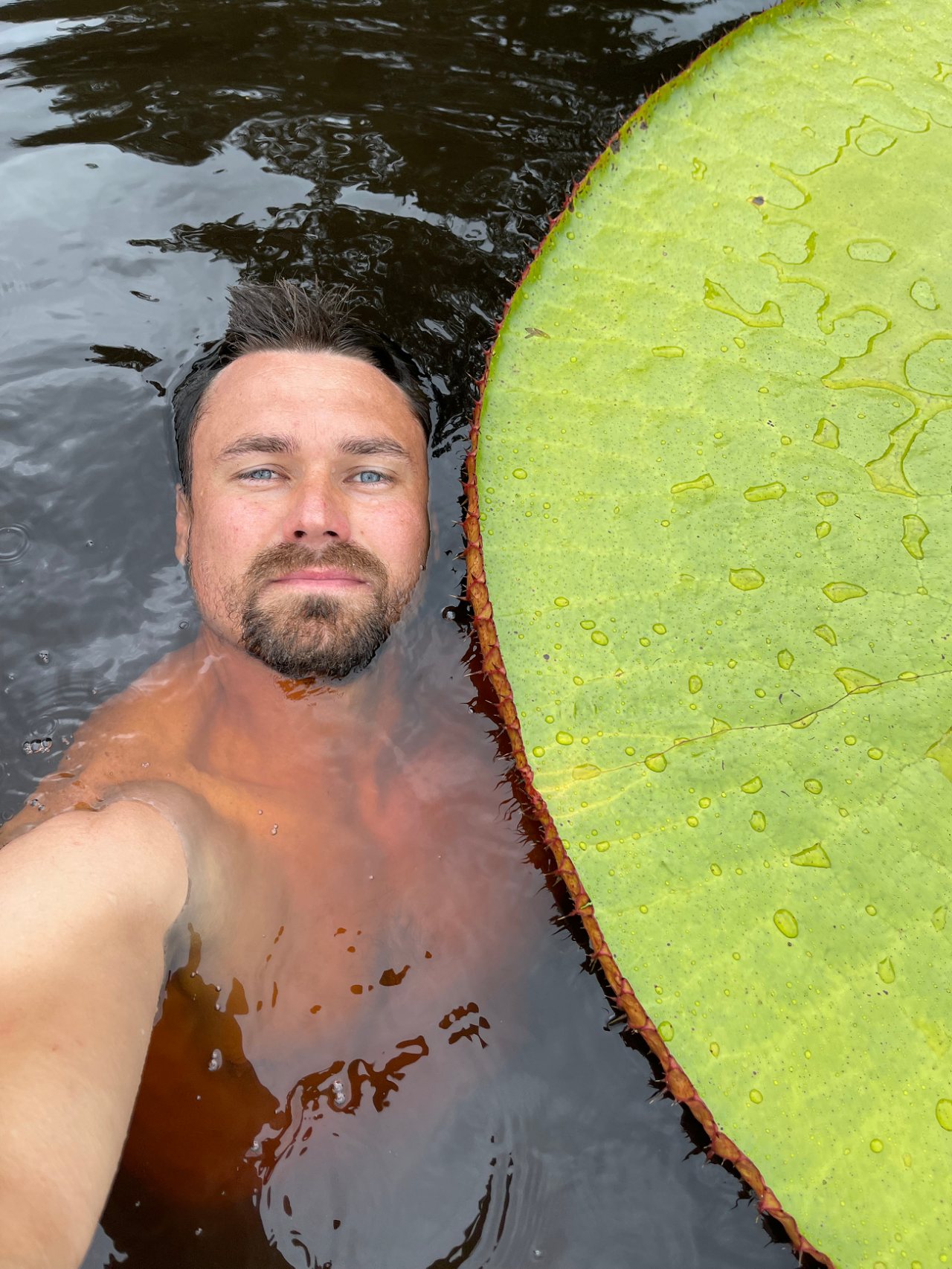 Jeremiah Harris taking a selfie while swimming in dark water beside an enormous, bright green giant waterlily leaf with a raised reddish rim in Colombia's Inírida River region.